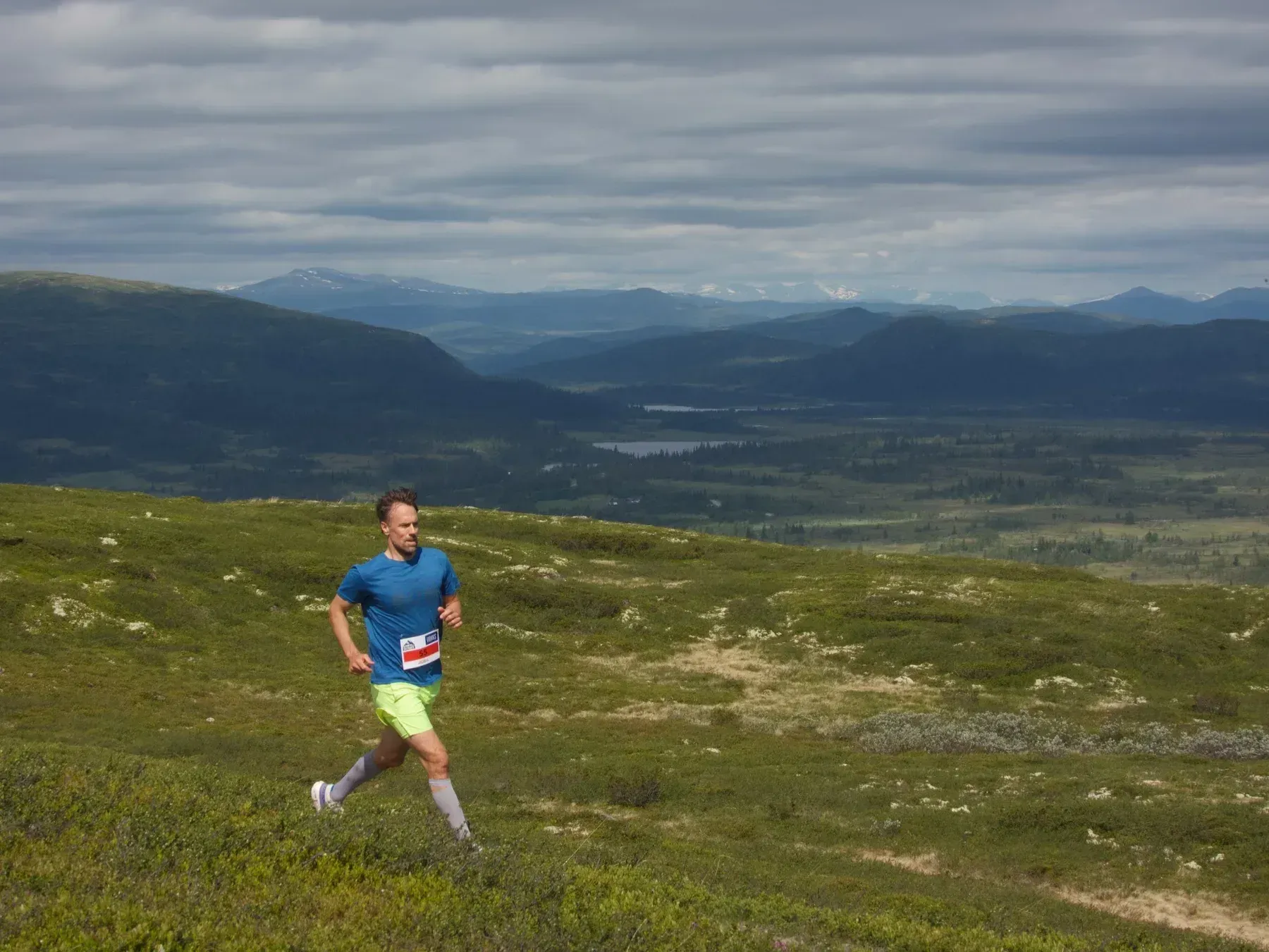 Løper i blått og grønt på fjellsti under skyer.