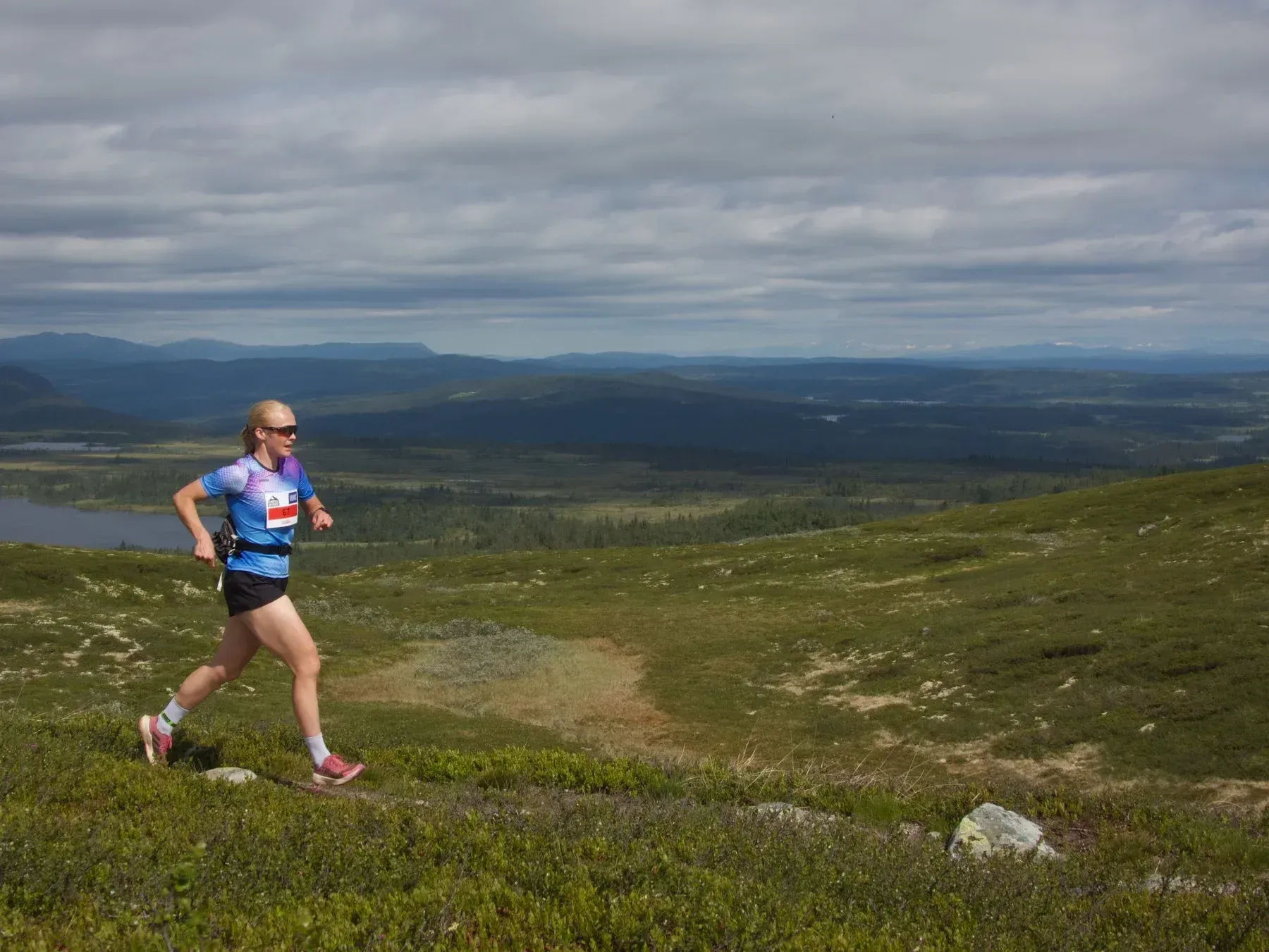 Løper med blå trøye og rød vest i åpent fjellterreng.