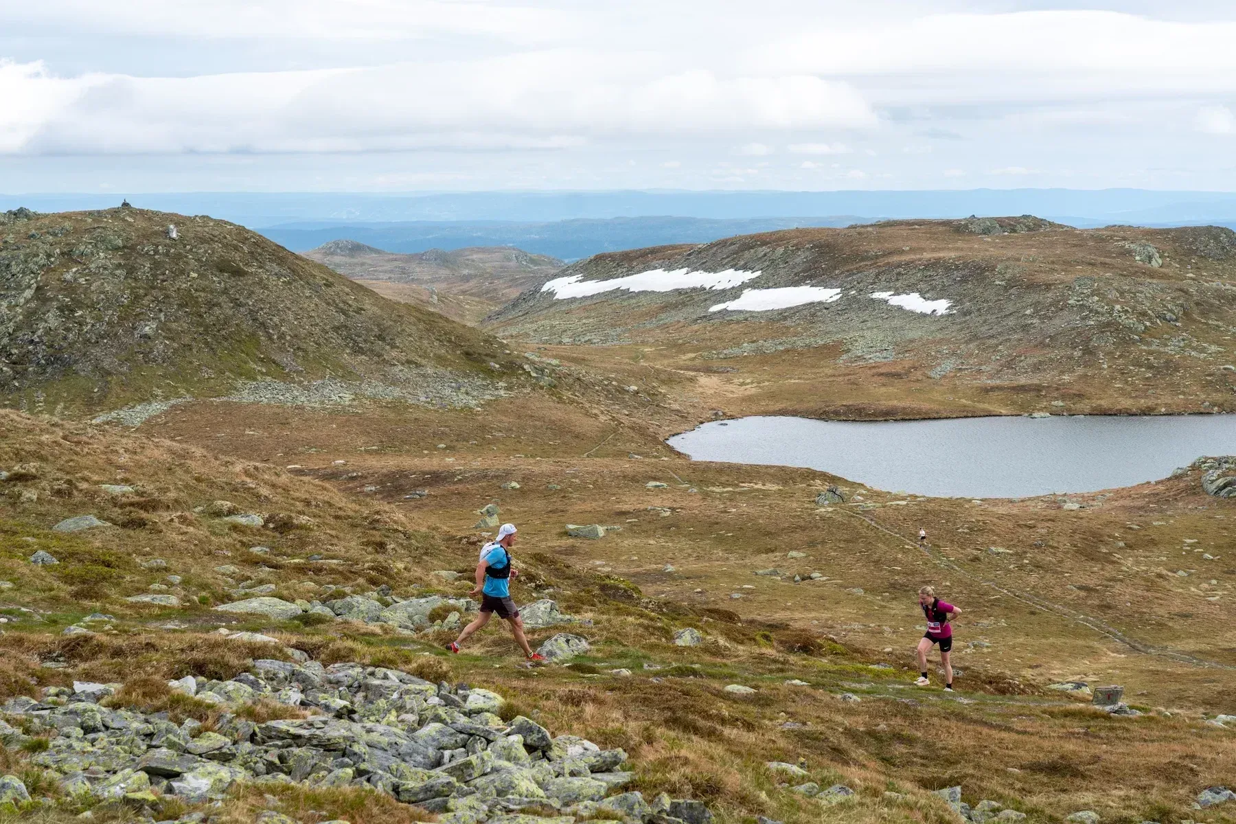 Løpere på fjellsti med utsikt mot et vann og snøflekker